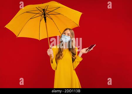 Une fille pensive dans un masque de protection sur son visage, dans une robe jaune et avec un parapluie dans ses mains, utilise un téléphone portable Banque D'Images