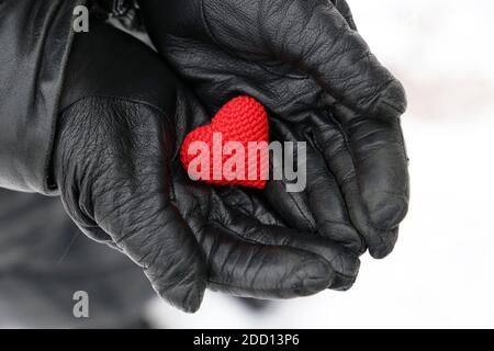 Coeur tricoté rouge dans les mains des femmes en gants de cuir noir sur fond de neige. Concept d'amour romantique, Saint-Valentin, soins de santé en hiver Banque D'Images