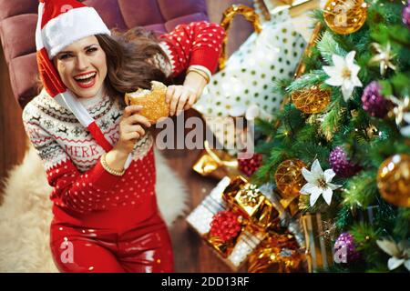 Joyeux Noël. Vue supérieure de la jeune femme en chandail rouge et chapeau de père Noël près de l'arbre de Noël et des boîtes-cadeaux manger du hamburger. Banque D'Images