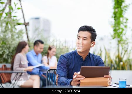 Portrait d'un jeune entrepreneur vietnamien pensif assis à une table de café avec table numérique et vue sur l'extérieur Banque D'Images