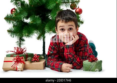 Portrait isolé sur blanc. Un garçon dans la chemise rouge se trouve sous un arbre de Noël, à côté de boîtes avec des cadeaux. Banque D'Images