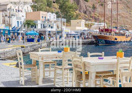 Chaises et tables d'une taverne grecque traditionnelle, restaurant au quai du petit port de bateau à voile flou et réveil touristique et boutiques. Santorin Banque D'Images