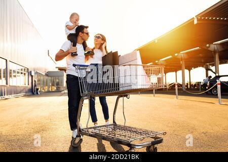 Une famille heureuse avec un enfant avec des sacs et un chariot dans la rue sur le fond d'un centre commercial, des magasins Banque D'Images