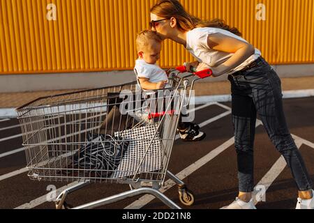 Belle jeune femme heureuse dans le shopping de lunettes de soleil avec petit enfant, chariot et sacs de shopping, à l'extérieur Banque D'Images