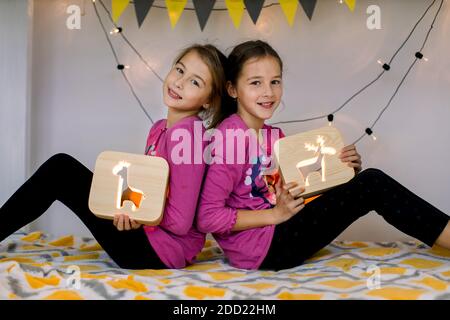 Heureux deux jolies sœurs filles de 10 ans souriantes, dans la chambre de l'enfant sur un lit superposé décoré, assis dos à dos et tenant des lampes de nuit en bois avec Banque D'Images