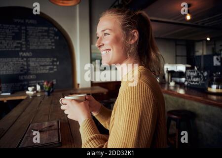 Souriante jeune belle femme tenant une tasse de café dans le café regardant loin Banque D'Images