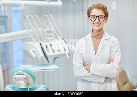 Portrait du jeune dentiste en manteau blanc debout avec les bras se croise en souriant à la caméra tout en se tenant à la clinique dentaire Banque D'Images