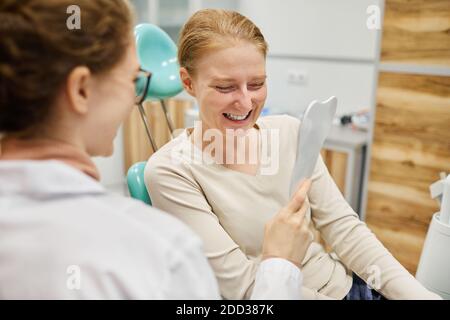 Jeune femme regardant le miroir elle est heureuse de sa nouvelle souriez en vous asseyant sur une chaise avec un dentiste à proximité Banque D'Images