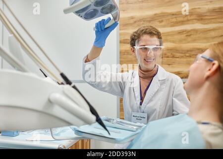Jeune dentiste en lunettes de protection souriant au patient pendant le traitement dentaire procédure à la clinique Banque D'Images