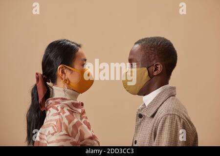 Vue latérale d'un jeune couple multiethnique dans des masques de protection l'un à l'autre isolé sur fond beige Banque D'Images