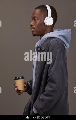 Vue latérale de l'homme africain dans des vêtements décontractés à l'écoute musique dans les écouteurs et café sur fond gris Banque D'Images