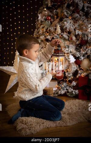 Petit garçon tenant une lanterne blanche dans sa main et assis près de l'arbre de Noël, bonne année Banque D'Images