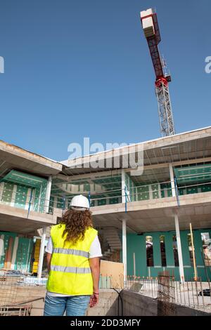 Vue arrière d'une jeune femme architecte portant un néon une veste de sécurité et un casque de sécurité blanc tout en regardant vers un chantier de construction Banque D'Images