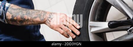vue rognée du technicien tatoué touchant la roue de voiture dans le garage, bannière Banque D'Images