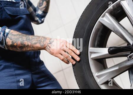 vue partielle du technicien tatoué touchant la roue de voiture dans l'atelier sur un arrière-plan flou Banque D'Images