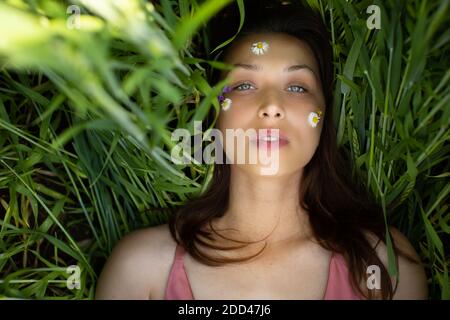 belle femme se trouve dans le blé vert avec des fleurs sauvages et marguerites collées sur le visage Banque D'Images