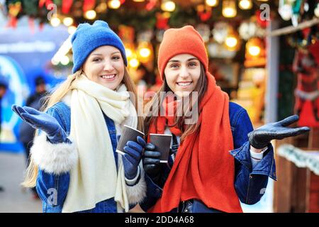 Deux jeunes filles en chapeaux et foulards rouges et bleus marchent autour du marché de Noël, boivent du café, parlent et sourient. Banque D'Images