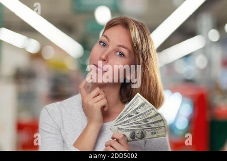 Finance et banque. Une jeune blonde tient une wad de dollars dans ses mains et sourit doucement. En arrière-plan se trouve un atelier flou. Banque D'Images