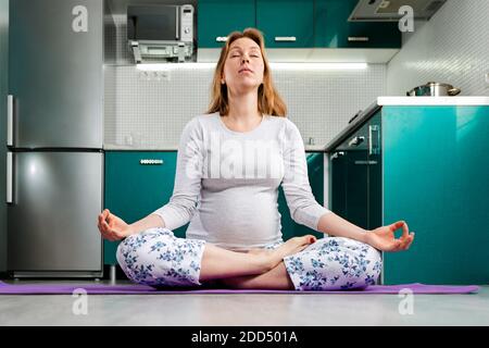 Auto-isolement et formation à domicile.UNE femme enceinte de race blanche fait du yoga et médite dans la position de Lotus. La cuisine est en arrière-plan. Concept o Banque D'Images