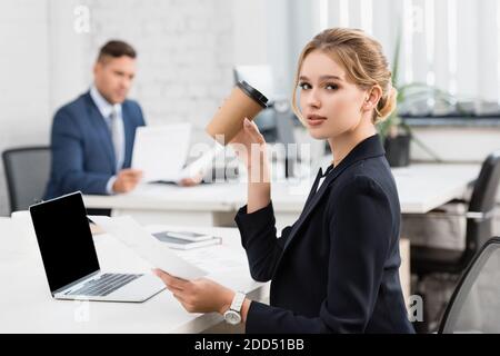 Femme d'affaires avec gobelet en papier regardant l'appareil photo, tout en tenant la feuille de papier sur le lieu de travail sur un arrière-plan flou Banque D'Images