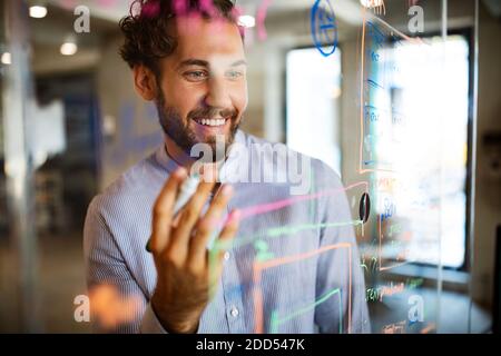 Jeune homme travaillant avec des bases de données et des diagrammes, écrivant des idées sur le mur de verre de bureau Banque D'Images