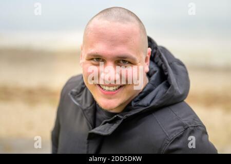 Jeune homme sympathique avec une tête rasée et un chaud souriez en vous penchant vers l'appareil photo pendant qu'il marche sur une plage par un froid jour d'automne Banque D'Images