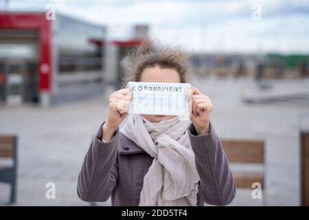 Portrait jeune femme européenne tenant un masque médical jetable de protection avec le texte manuscrit du coronavirus couvre le visage, dans la rue de ville. Concept de Banque D'Images