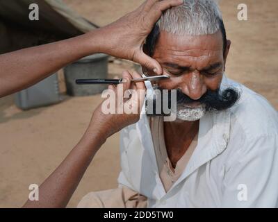 Un homme avec une longue moustache reçoit un rasage et une coupe de cheveux par le barbier de rue à l'aide d'un rasoir à gorge coupée à Pushkar, Rajasthan, Inde. Banque D'Images