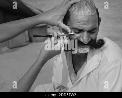 Un homme avec une longue moustache reçoit un rasage et une coupe de cheveux par le barbier de rue à l'aide d'un rasoir à gorge coupée à Pushkar, Rajasthan, Inde. Banque D'Images