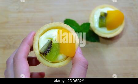 Une main tenant une mini tarte aux fruits, avec une autre sur la table dans le fond flou. Banque D'Images