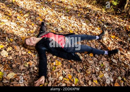 Belle femme en train de rire, couchée sur un tapis de feuilles mortes et faisant de l'ange dans un feuillage d'automne luxuriant dans la forêt. Détente et amusement. Banque D'Images