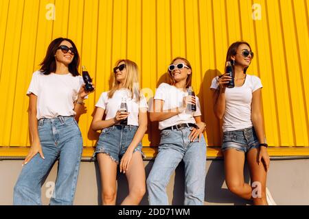 Souriante et élégante, les jeunes femmes sont en lunettes de soleil avec des boissons debout mur jaune dans la ville sur le soleil Banque D'Images