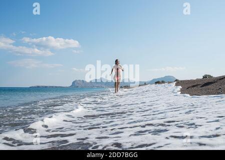 Jeune femme en bikini rose courant à côté de la belle plage de galets avec des vagues mousseuse entrant. Banque D'Images