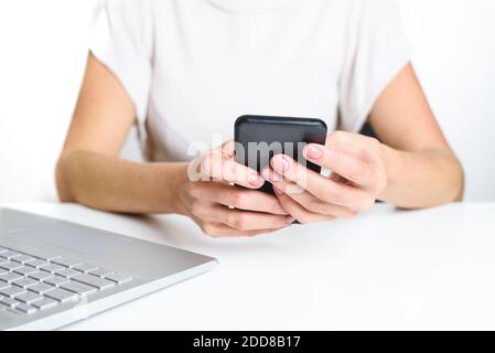 les mains d'une jeune femme caucasienne méconnaissable tenant un smartphone sur le dessus d'un bureau blanc avec un ordinateur portable moderne à proximité. fond blanc et sele Banque D'Images