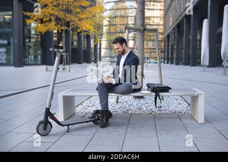 Homme d'affaires souriant utilisant un ordinateur portable tout en étant assis en poussant sur le scooter banc en ville Banque D'Images