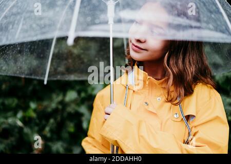 Fille en imperméable regardant loin en se tenant sous le parapluie à l'extérieur Banque D'Images