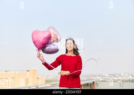 Femme mûre tenant un ballon en forme de coeur tout en étant debout sur le bâtiment terrasse au ciel clair Banque D'Images