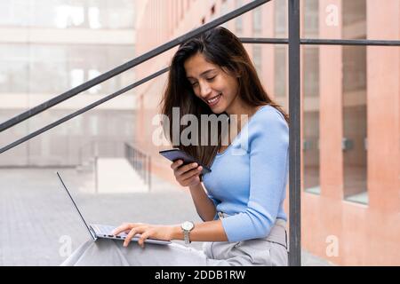 Jeune femme d'affaires souriante utilisant un smartphone tout en étant assise avec un ordinateur portable bâtiment de bureau extérieur Banque D'Images