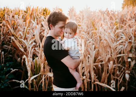 Père transportant un bébé garçon tout en se tenant contre le terrain pendant le coucher du soleil Banque D'Images