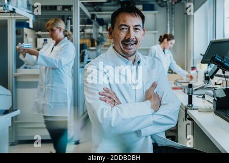 Homme scientifique assis avec les bras croisés par microscope pendant que collègue travailler en arrière-plan au laboratoire Banque D'Images