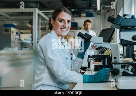 Femme souriante analysant le cerveau humain glisser au microscope tout en étant assise avec des scientifiques en laboratoire Banque D'Images