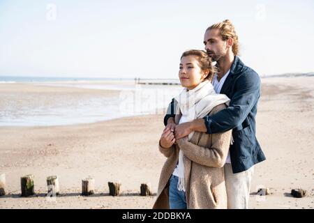 Jeune couple qui regarde loin tout en embrassant à la plage Banque D'Images