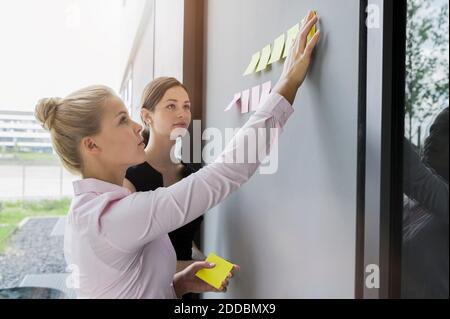 Femme d'affaires qui colle des notes adhésives sur le mur tout en discutant avec un collègue au bureau Banque D'Images