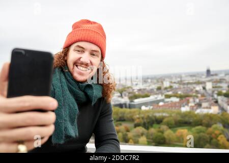 Homme souriant tout en prenant le selfie à travers le smartphone dans le bâtiment terrasse contre le ciel Banque D'Images