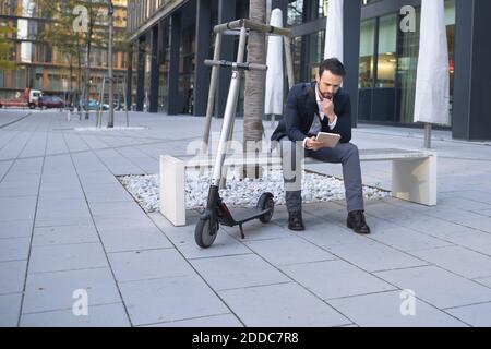 Homme d'affaires utilisant une tablette numérique tout en étant assis à bord d'un e-scooter sur un banc en ville Banque D'Images