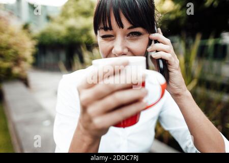 Femme d'affaires souriante tenant une tasse de café tout en parlant sur un téléphone portable Banque D'Images