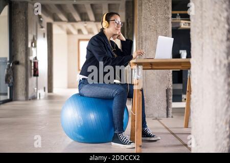 Femme d'affaires portant un casque avec un ordinateur portable tout en étant assise sur un ballon de fitness au bureau Banque D'Images