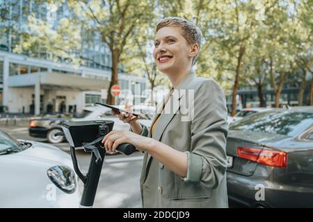 Femme déverrouillant un scooter électrique avec un smartphone debout sur la rue en ville Banque D'Images