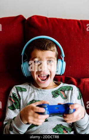 Joyeux garçon portant un casque tout en jouant à un jeu vidéo sur un canapé à la maison Banque D'Images