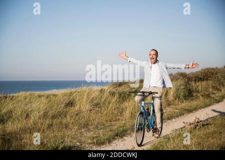 Homme mûr heureux avec les bras débordés à vélo sur la plage contre le ciel dégagé Banque D'Images